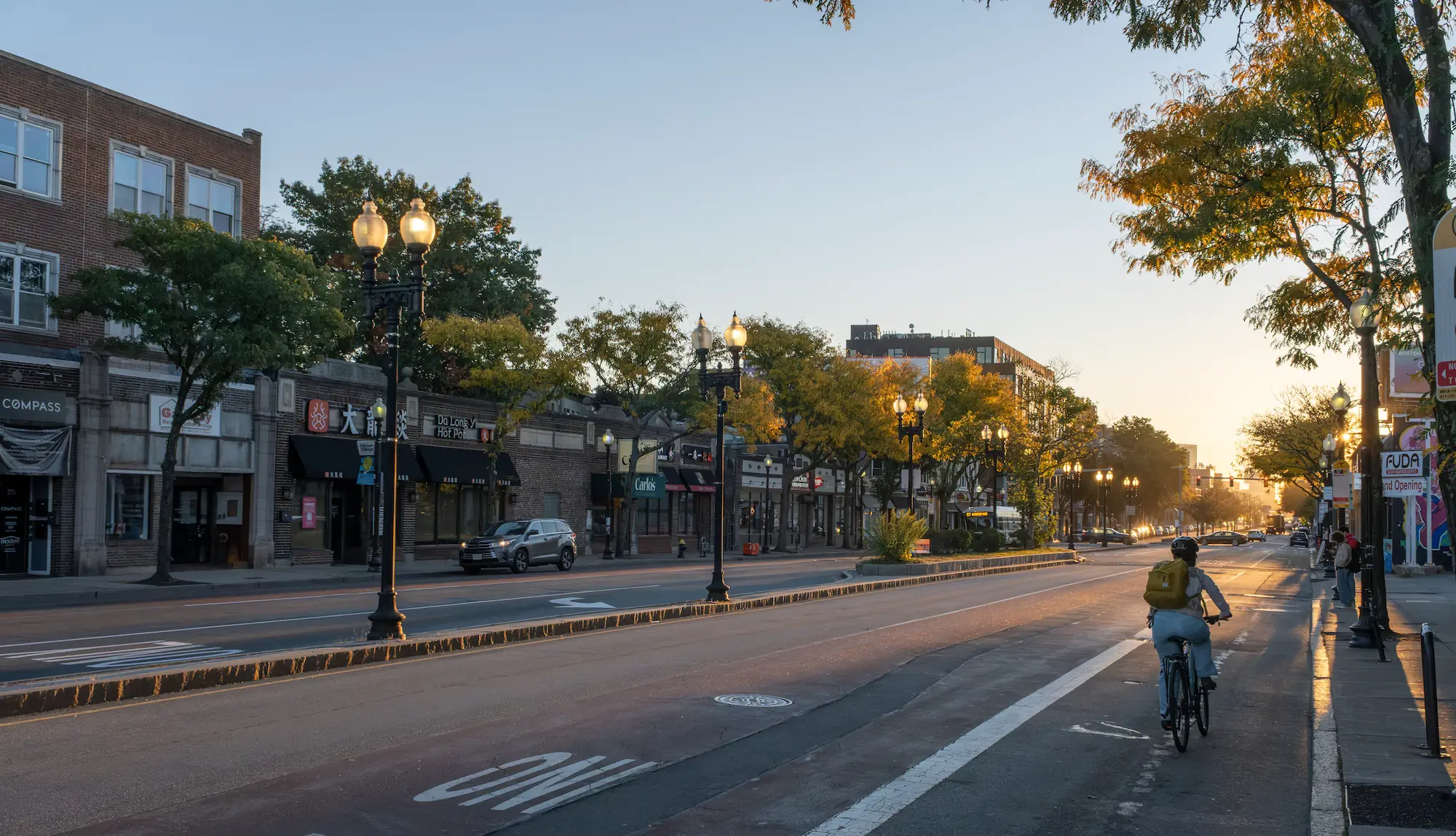 Brighton Avenue in Allston at golden hour, a cyclist in the bike lane and lit lamp posts down the block