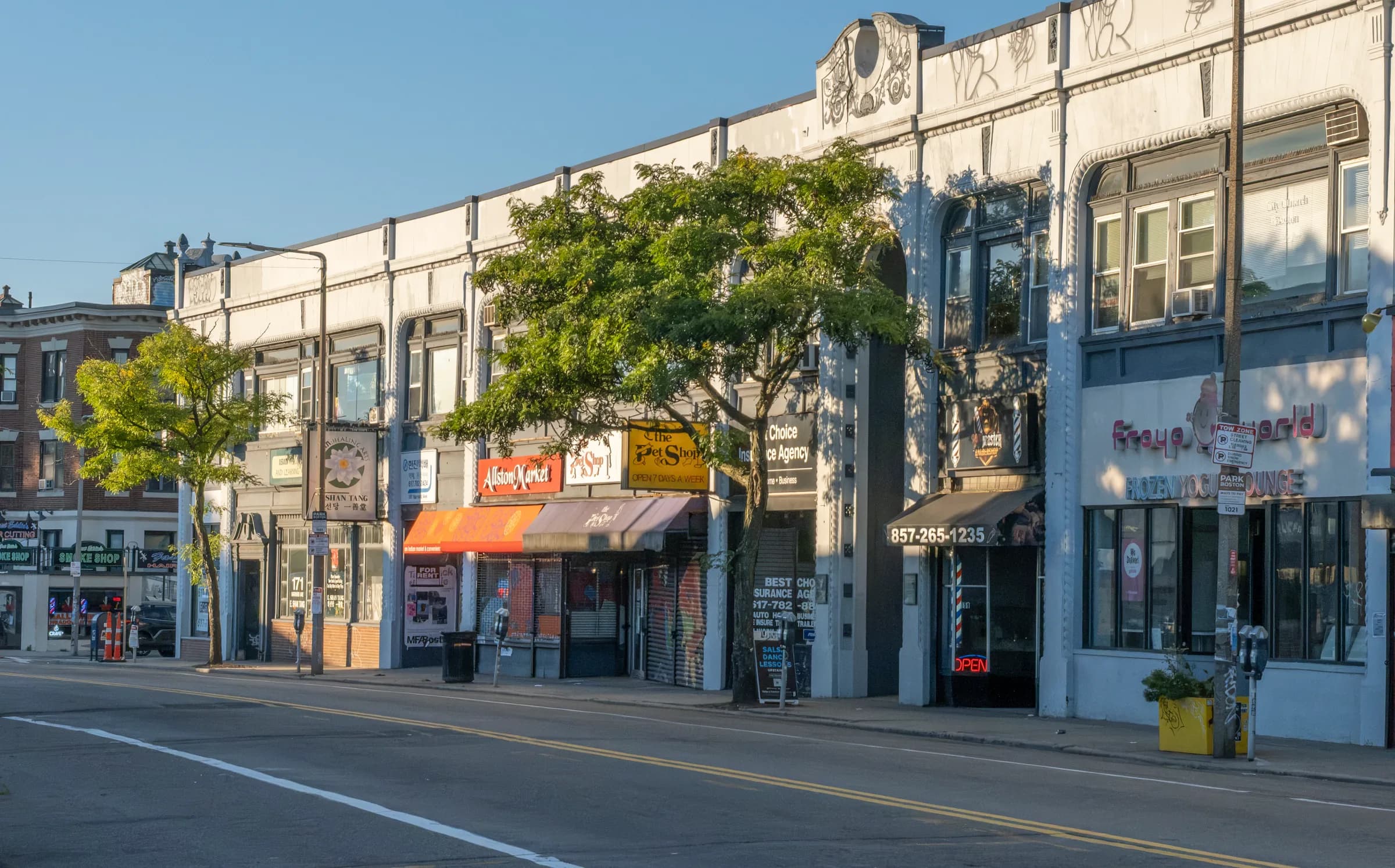 A row of Harvard Avenue storefronts in Allston in early light, awnings and signs under one long white facade
